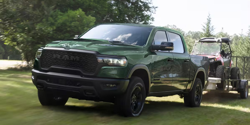 A 2025 RAM 1500 truck in green towing an ATV on a trailer through a grassy field.