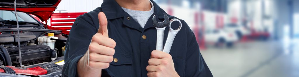 A mechanic giving a thumbs up while holding two wrenches, with a car engine in the background and a blurred workshop setting.