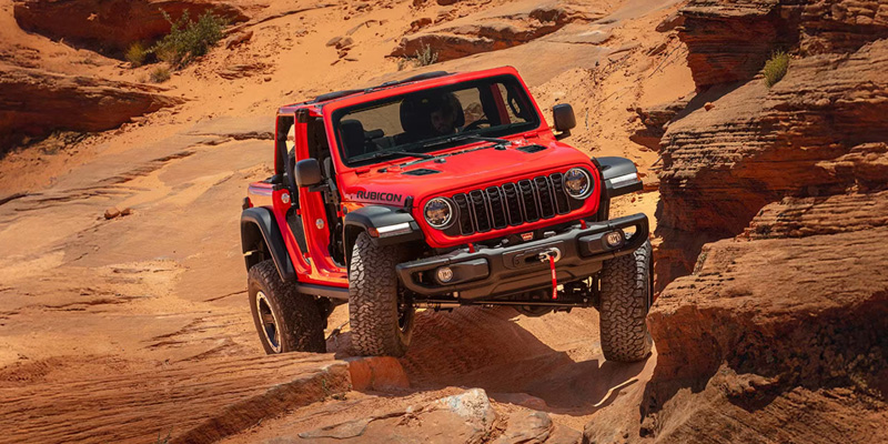 Red Jeep Wrangler driving through a rugged rocky canyon, navigating uneven terrain with off-road capability.