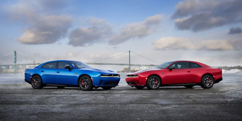 Side view of a blue and a red Dodge Charger facing each other on an open road near a bridge, showcasing sporty performance styling under a cloudy sky.