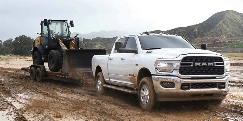 Ram pickup truck towing a travel trailer on an open road through a scenic landscape.